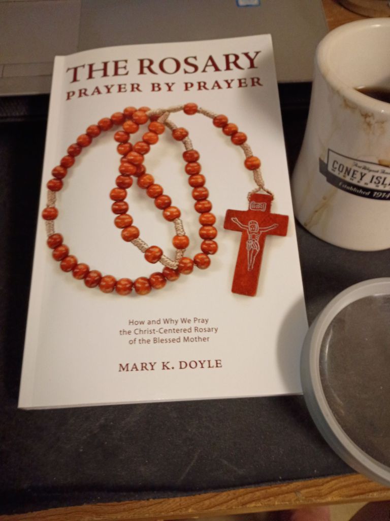 A close-up of the book 'The Rosary Prayer by Prayer' by Mary K. Doyle, featuring a wooden rosary with a crucifix and a cup of coffee beside it.